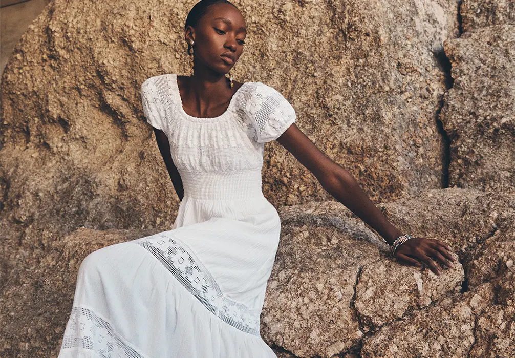 Model standing by rocks at the beach wearing the Floral Lace Cotton Milkmaid Maxi Dress