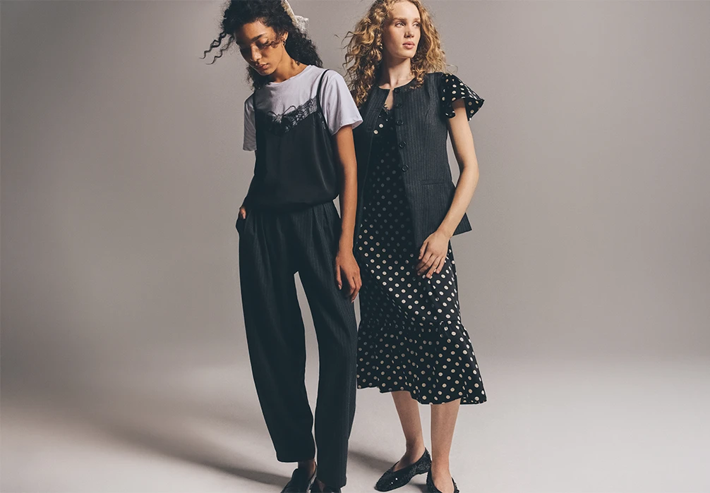 Two models standing wearing a polka dot dress with a waistocat, and a black lace cami top over a white tshirt