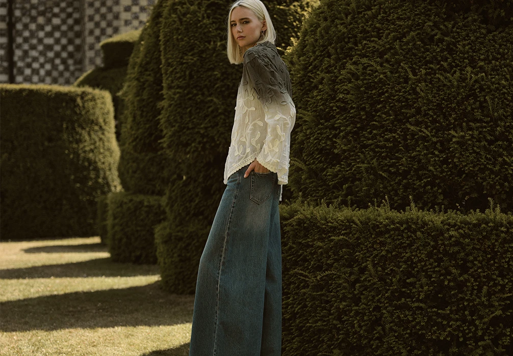 A girl standing in Jeans and a crochet top. Text on image: Cool and casual, explore timeless denim designed for off-duty days. Take your pick from our collection of jeans, from figure-showcasing skinny jeans and silhouette-skimming barrel jeans, to retro-cool flare jeans and wide-leg jeans. Versatile and easy to style, we've got a pair to fit and flatter you.
