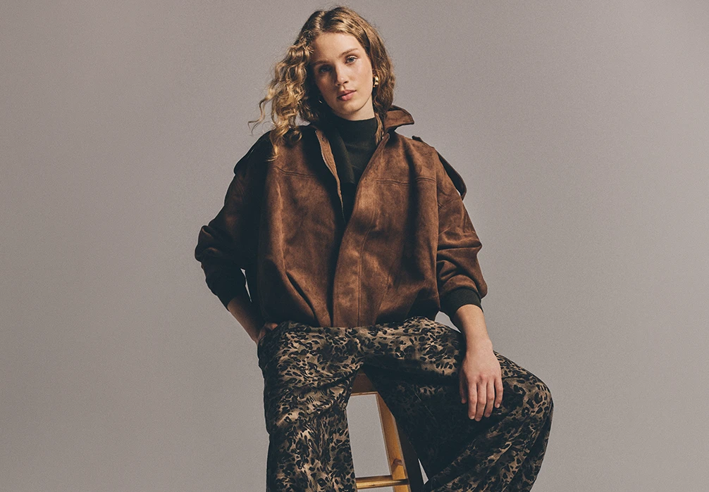 Model is sat wearing a brown suede bomber jacket with leopard print jeans
